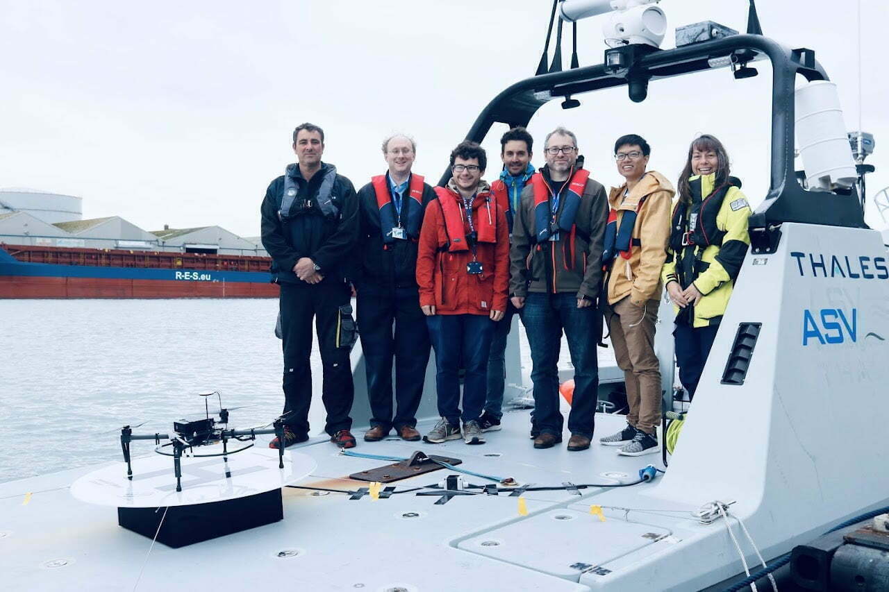 The Bumblebee team standing on a boat. A drone that is wirelessly charging sits nearby.